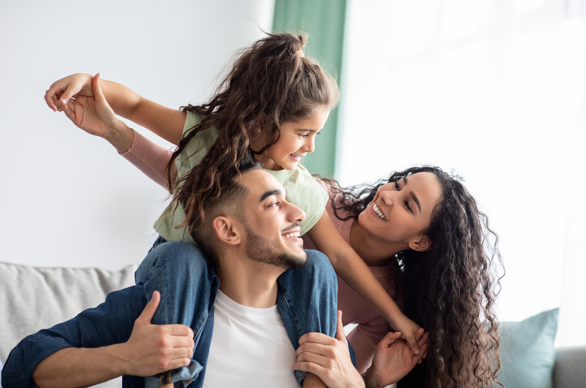 A joyful family moment featuring a father with his daughter on his shoulders, both smiling and looking at the mother, who is lovingly engaging with them.